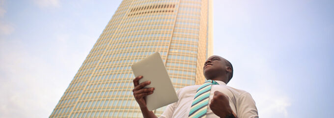 A professionally dressed young adult standing outside a large office building and celebrating while holding a job offer